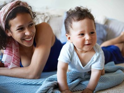 Childhood, physical development and childcare concept. Picture of attractive cheerful young mixed race female laughing, watching her adorable toddler child making first crawling movements on floor